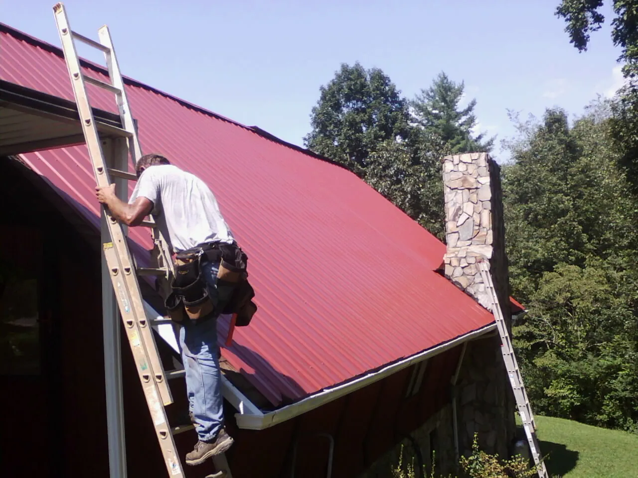 Roofer on a ladder beside a red metal roof and stone chimney — Soffit & Fascia Repair by Roofing Brothers in Church Hill